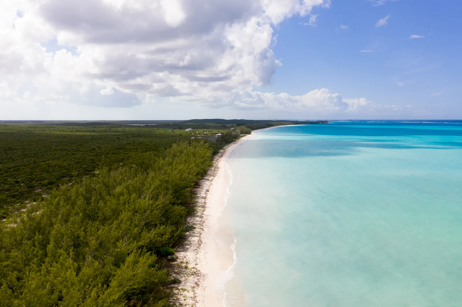 An empty white beach between green scrub and pale turquoise water, seen from the air