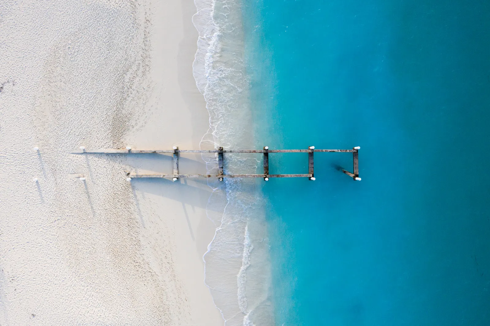 The posts of an old wooden pier on white sand and bright turquoise water, seen from straight above
